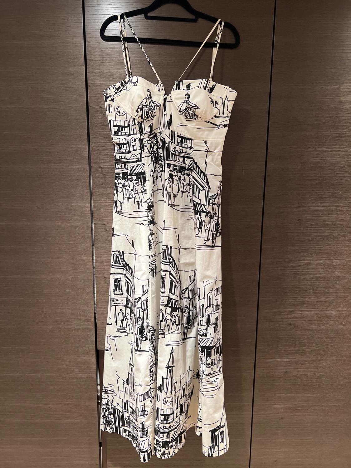 Long white strappy dress with a black cityscape print, hanging on a black hanger in front of a dark wooden wardrobe.