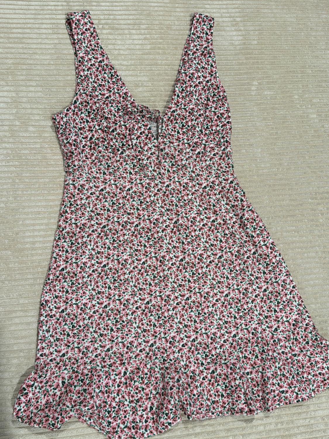 Short sleeveless dress with a pink floral print, V-neckline, and ruffled hem, set against a light background with a ribbed texture.