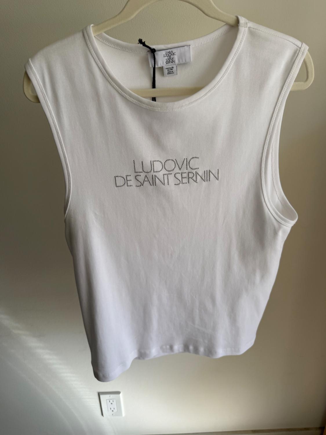 White sleeveless top hanging on a hanger, with the text "LUDOVIC DE SAINT SERNIN" on the front. Neutral background.