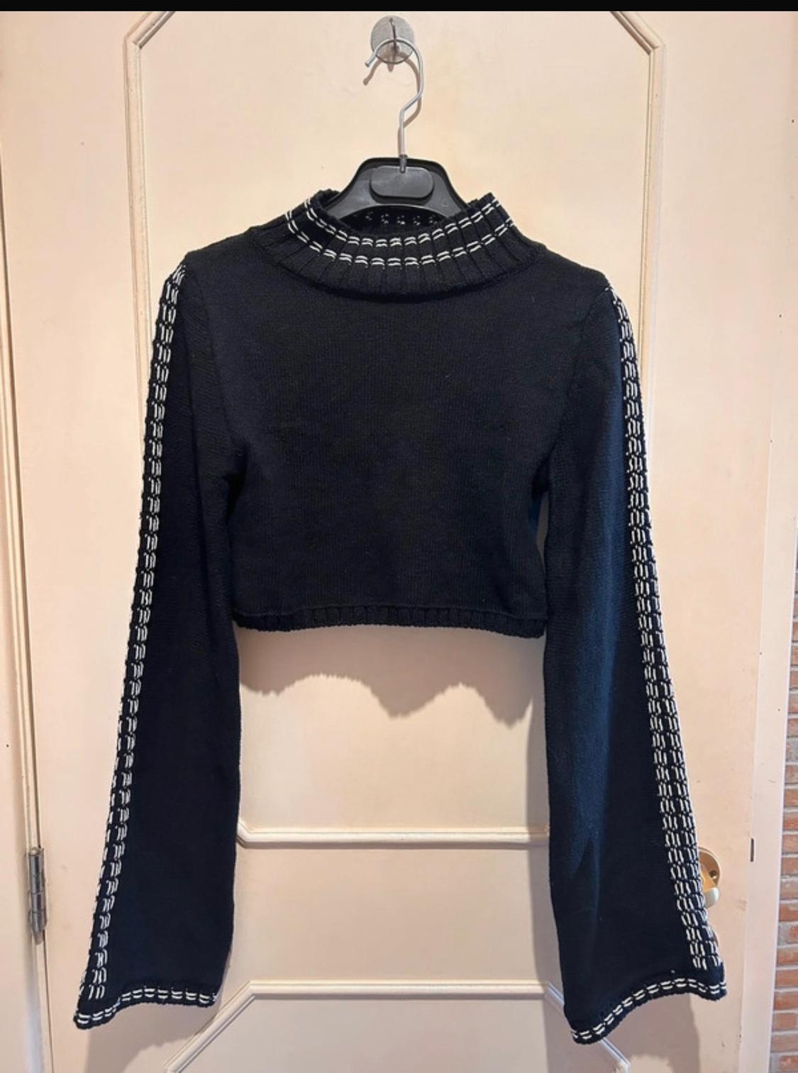 Black cropped jumper with a high neck and long sleeves. White knit details on the neck and sleeves. Hanging on a door.
