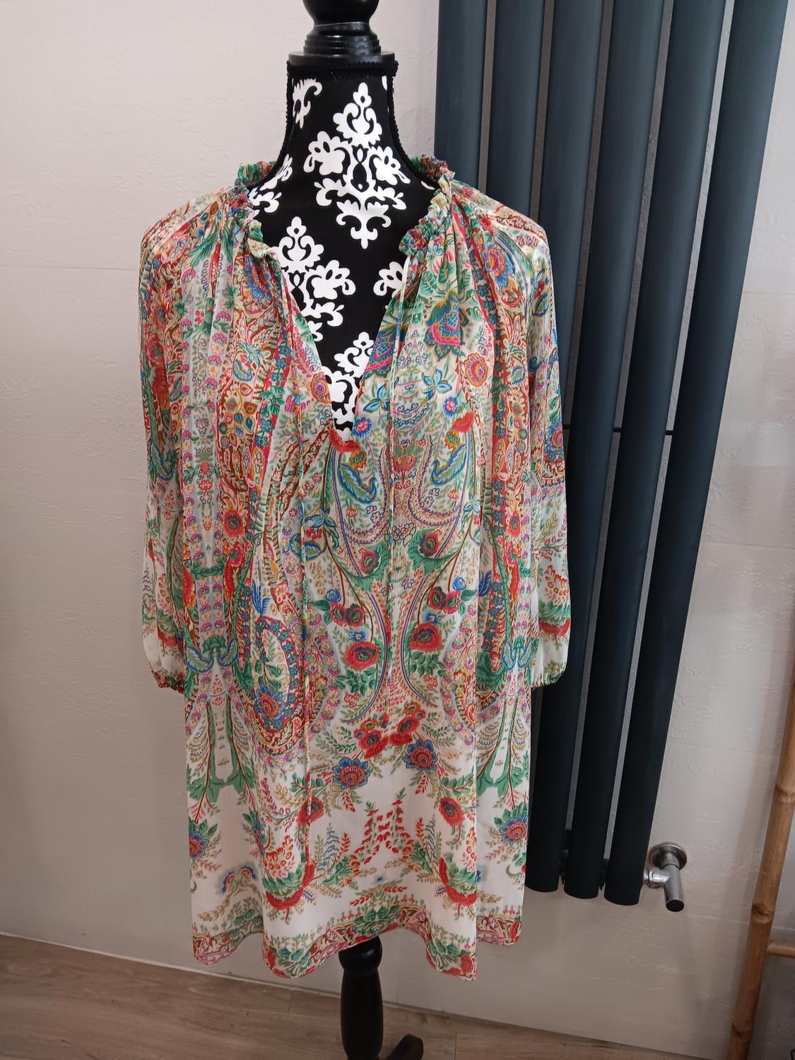 Colourful patterned dress with floral and paisley motifs on a black mannequin with a white design, dark curtains in the background.