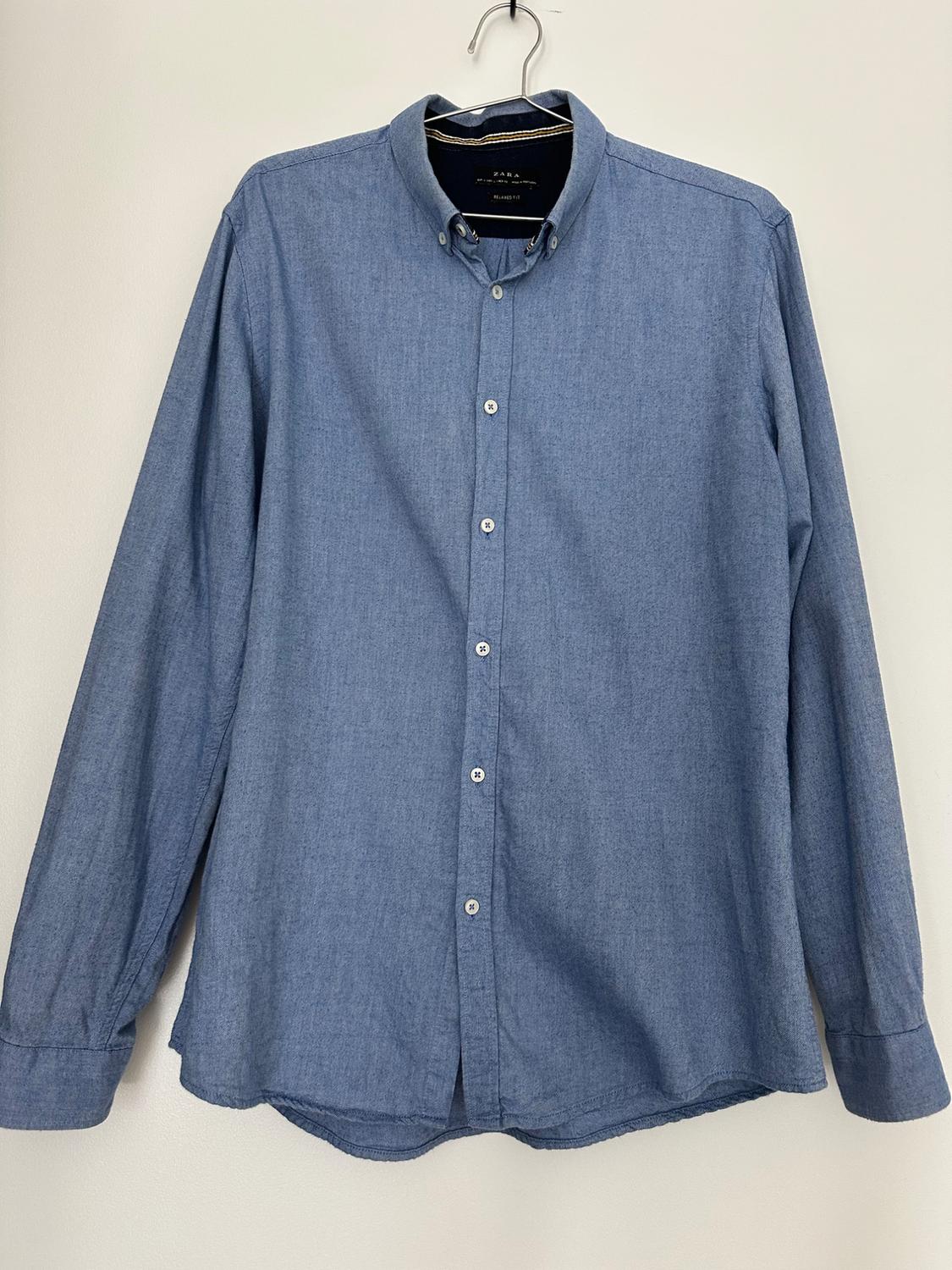 Blue long-sleeve shirt with white buttons, hanging on a hanger against a light background. Tag with the text ZARA RELAXED FIT.