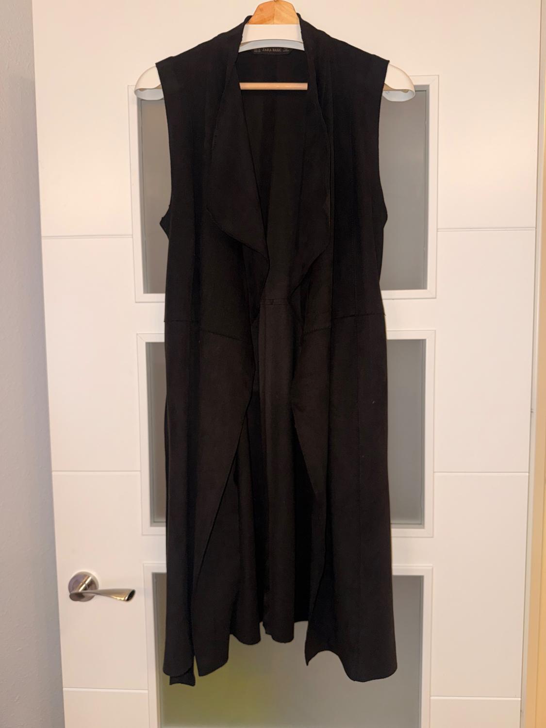 Long black sleeveless waistcoat hanging on a hanger over a white door with glass panels. Visible text: ZARA BASIC.