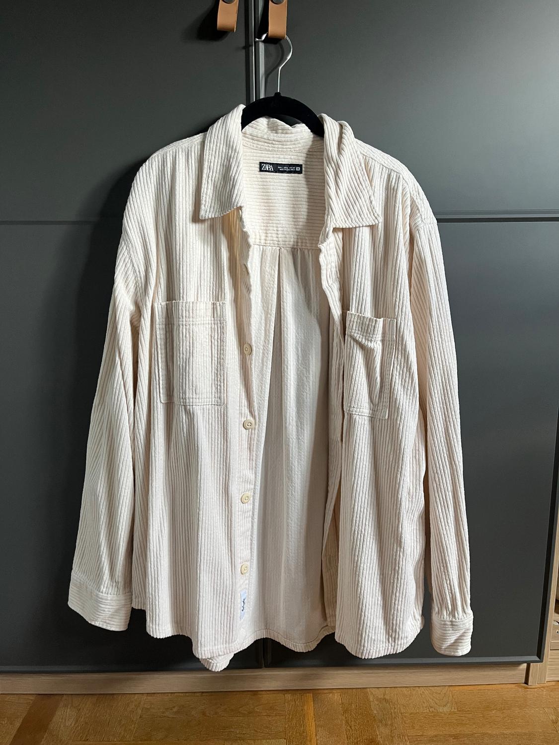 White vertical striped shirt hanging on a black hanger in front of a grey wardrobe. Visible tag: "Zara".