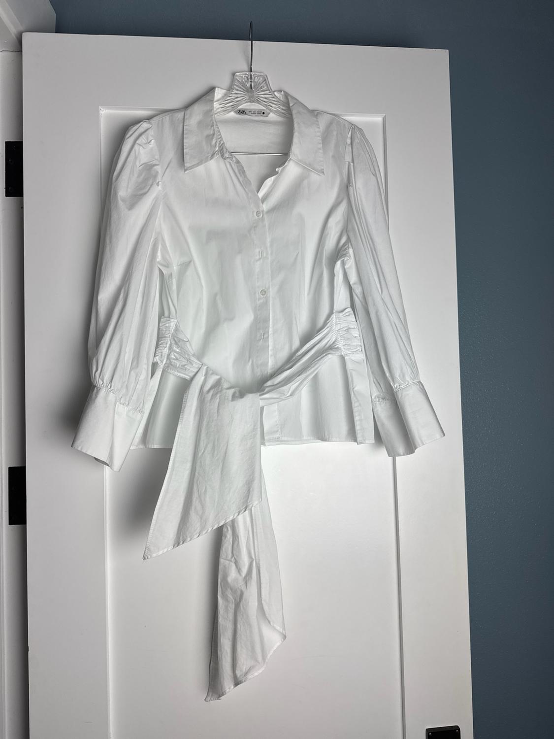 White ZARA button-up shirt with puff sleeves and a waist tie, hanging on a white door.