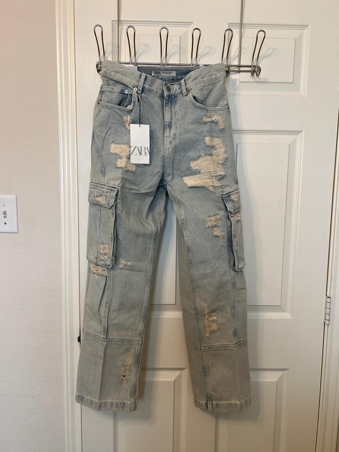 Light-blue ripped jeans with cargo pockets, hanging on a white door. Includes a tag bearing the text ZARA.