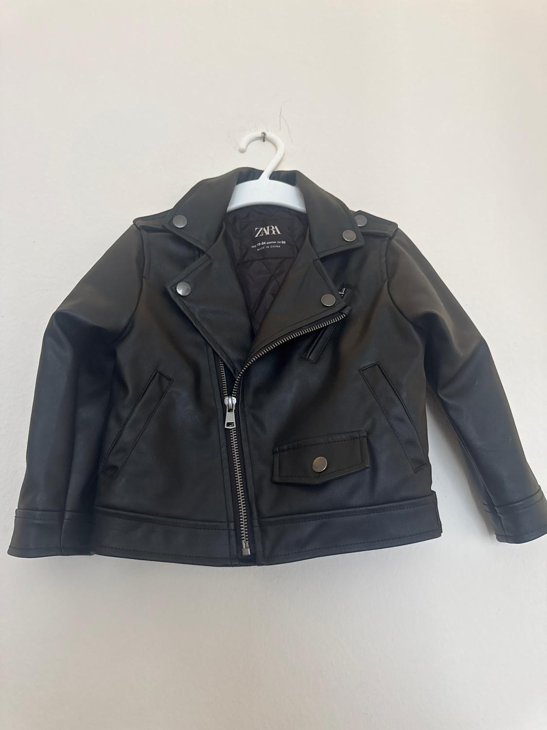 Black faux leather jacket for children hanging on a white hanger. Features zips and metal buttons. Visible brand: Zara.