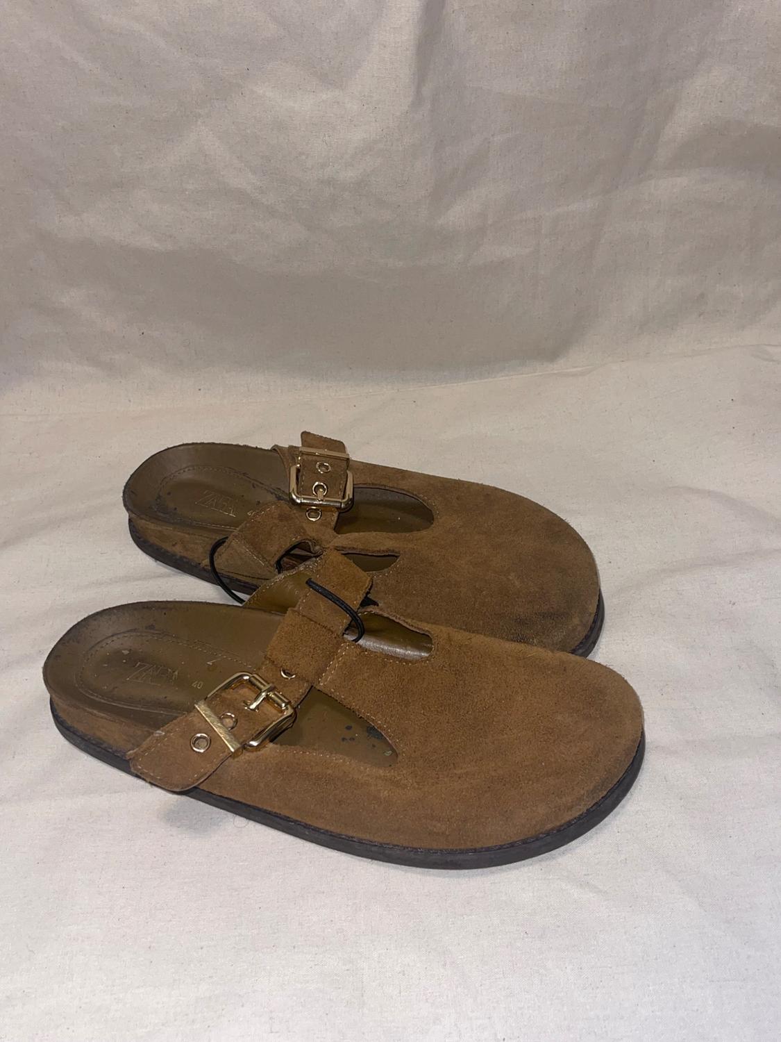 Pair of brown suede clogs with a gold buckle on a wrinkled light-colored fabric background. Visible text: ZARA 40.