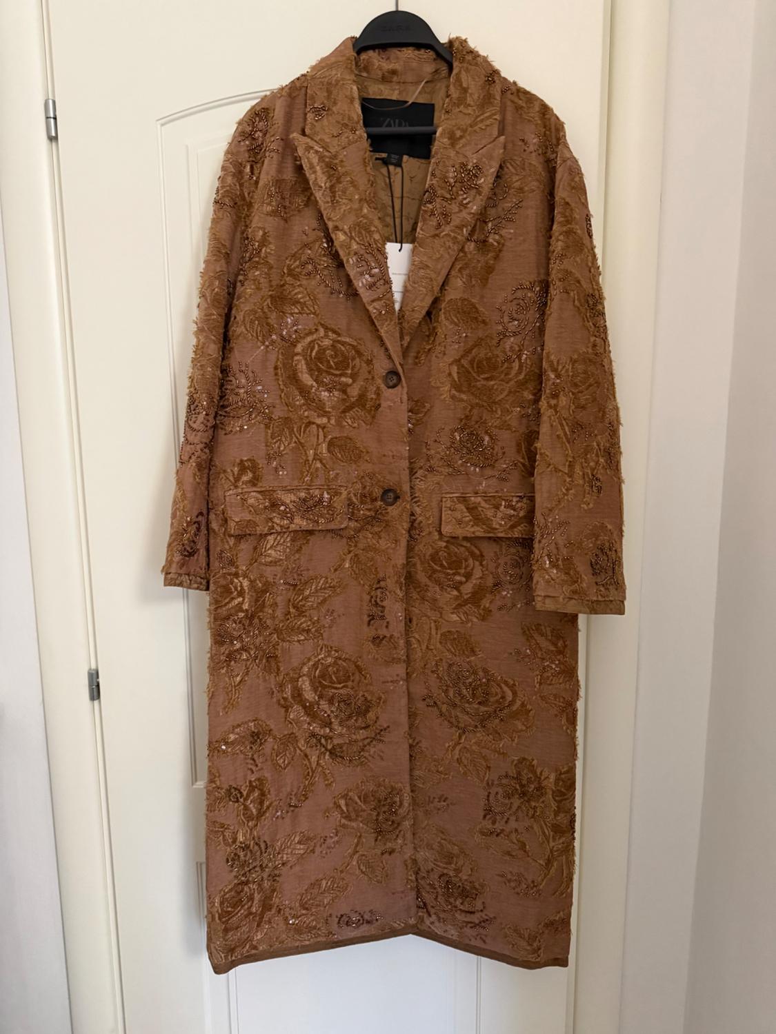 Long brown coat with floral embroidery hanging on a black hanger over a white door.