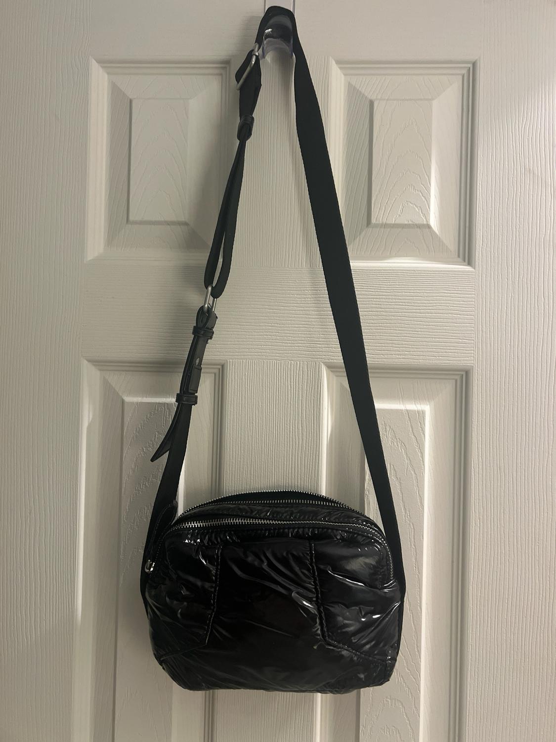 Black quilted, glossy crossbody bag hanging on a white door. Casual design with an adjustable strap and multiple zippers.