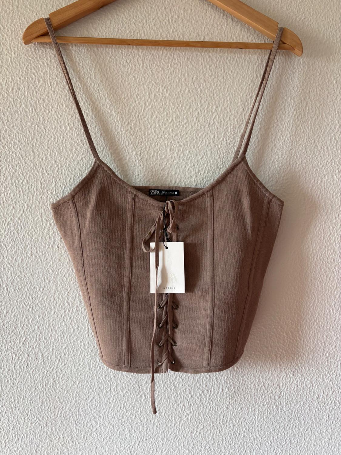 Brown camisole top with corset-style front lacing, hanging on a wooden hanger against a white wall. Visible text: ZARA LINGERIE.