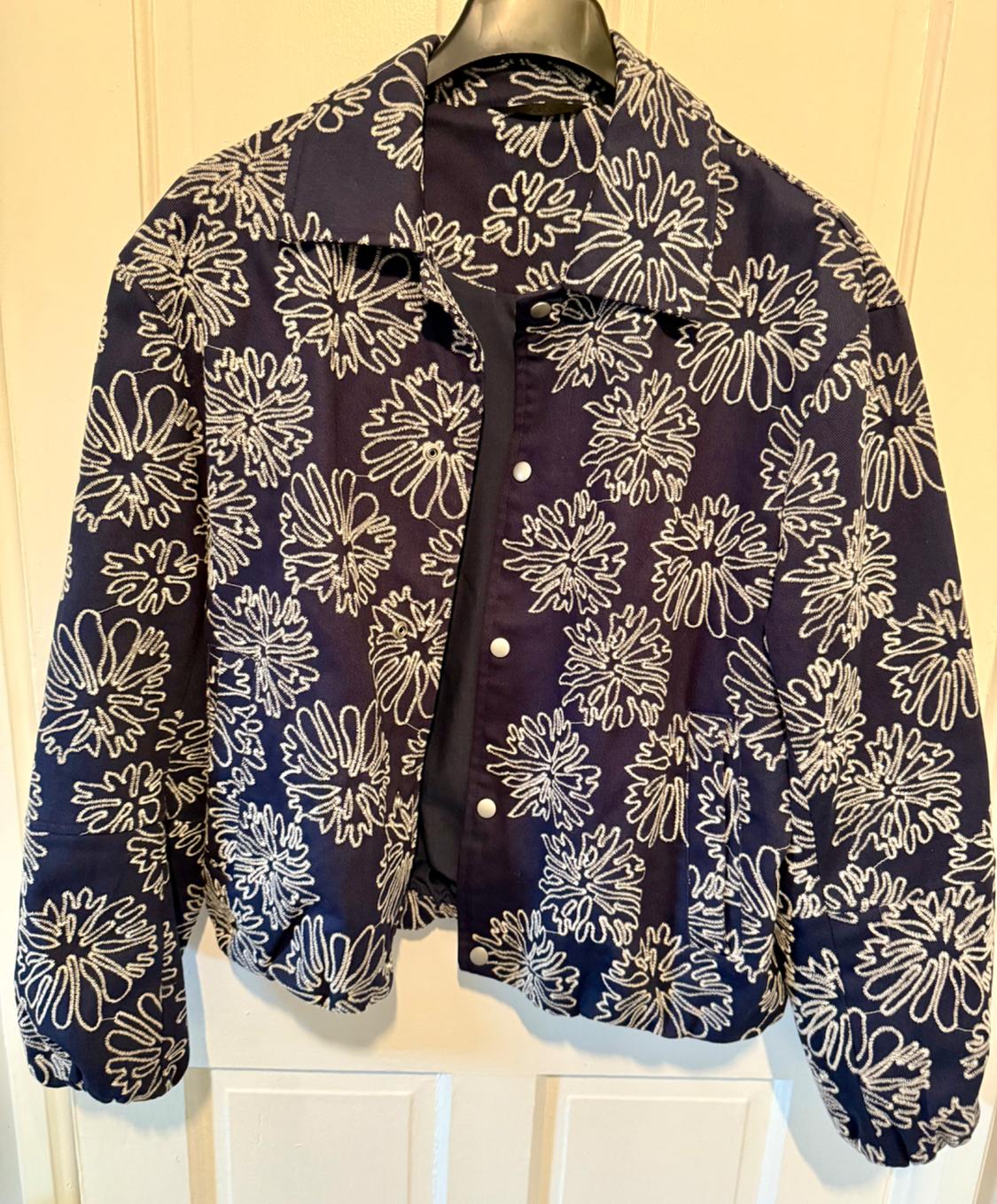 Blue jacket with white floral print, hanging on a hanger. Large floral design and visible front buttons.