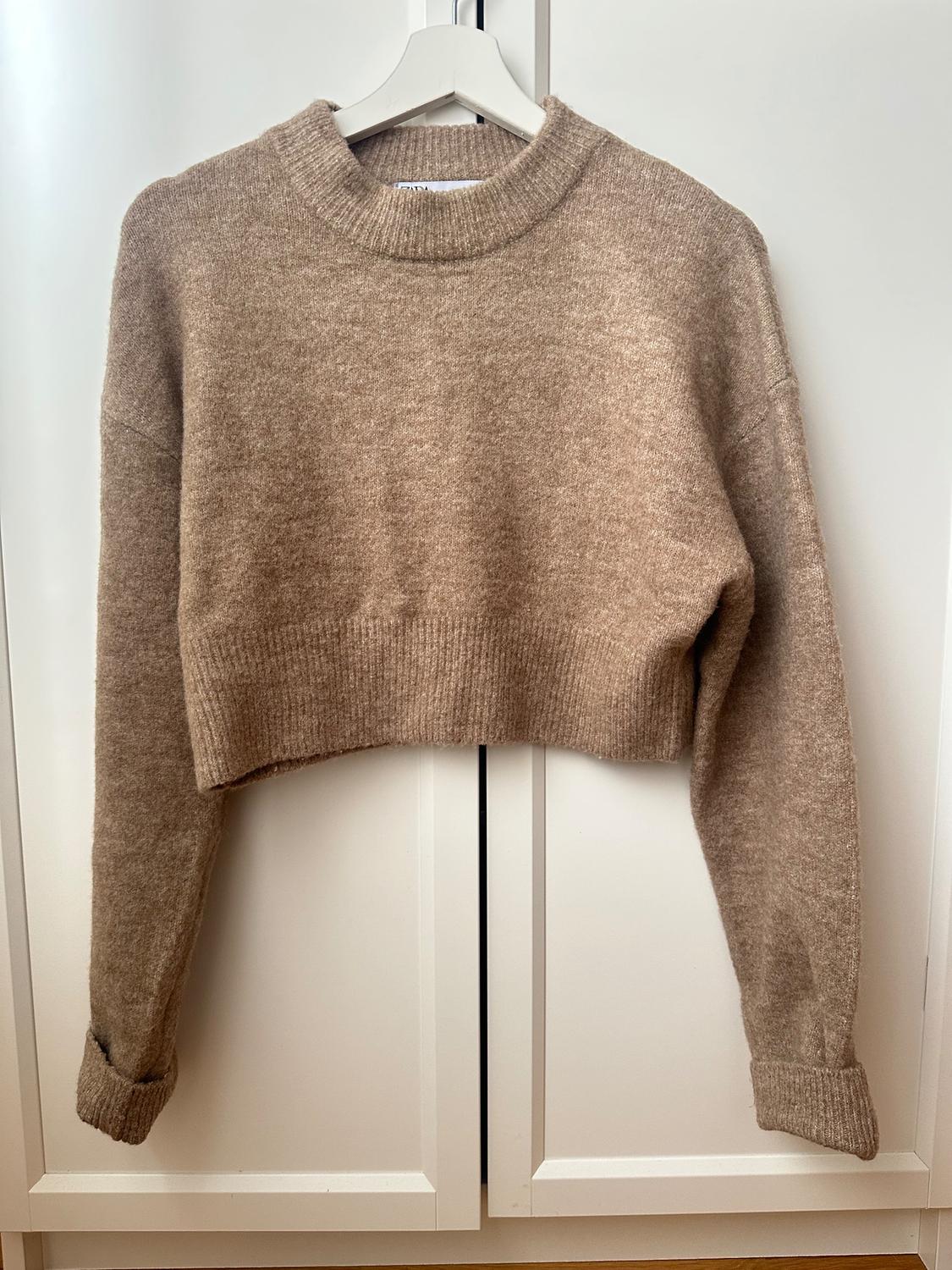 Brown knitted cropped jumper hanging on a white hanger in front of a white wardrobe. Long sleeves with folded cuffs.