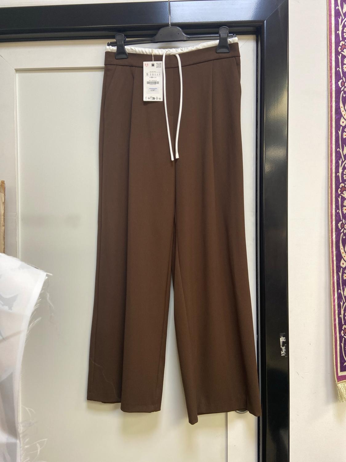 Brown wide-leg trousers with an elasticated waist and white drawstring, hanging on a hanger over a white door with a black frame.