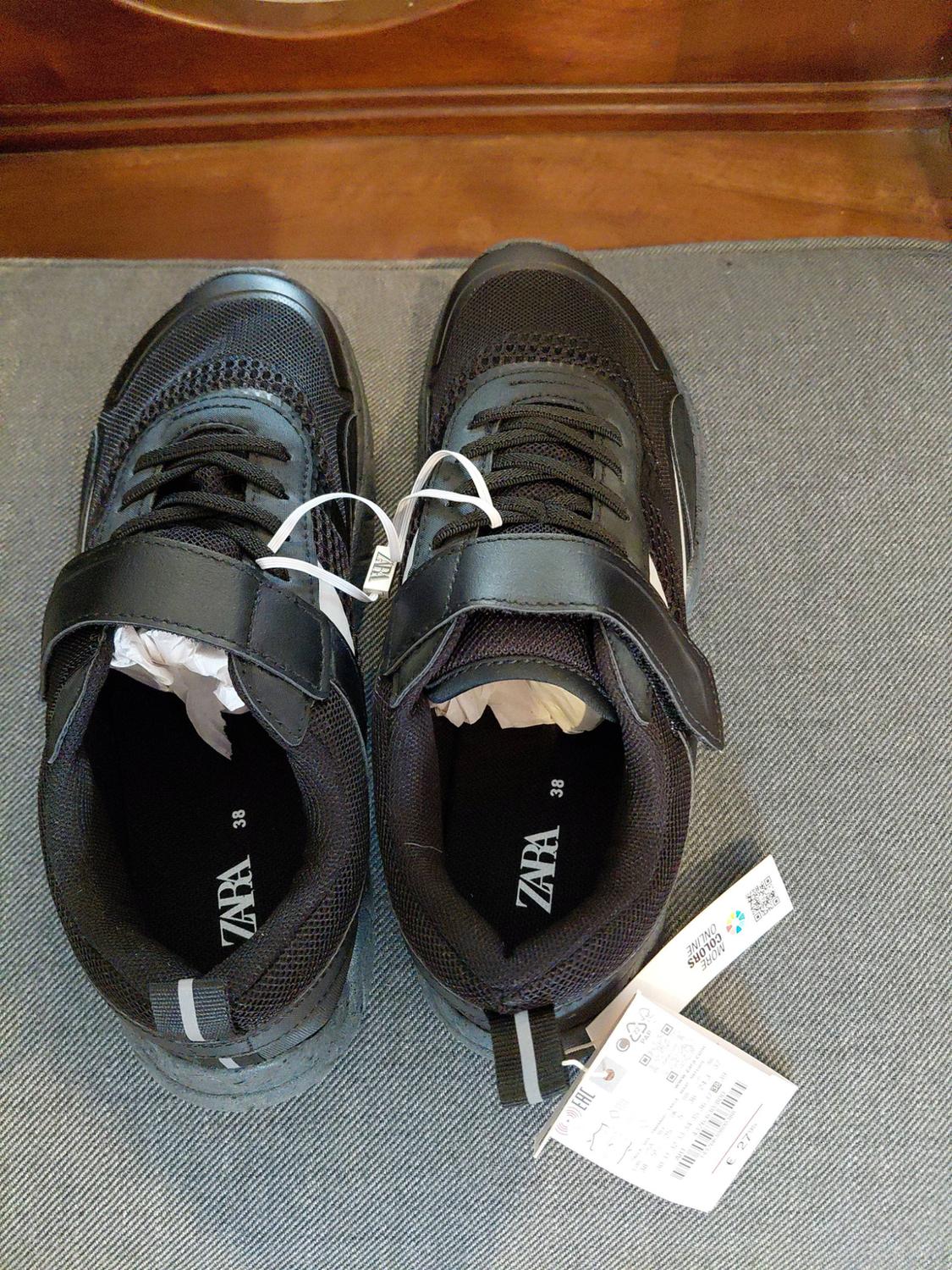 Black trainers with laces and labels. Text: ZARA, 38, MORE COLOURS ONLINE, €27.95, EAC. On a grey and wooden surface.
