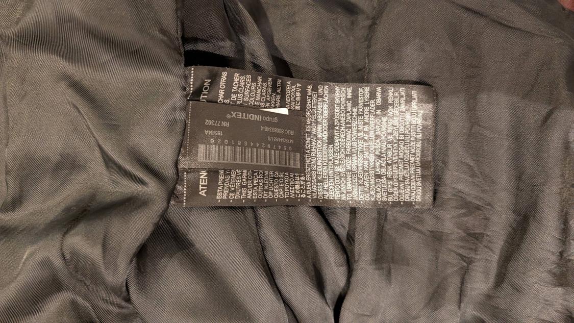 Close-up of a black label sewn into the dark lining of a garment. It contains multilingual text, "INDITEX group" and "RN 77302".