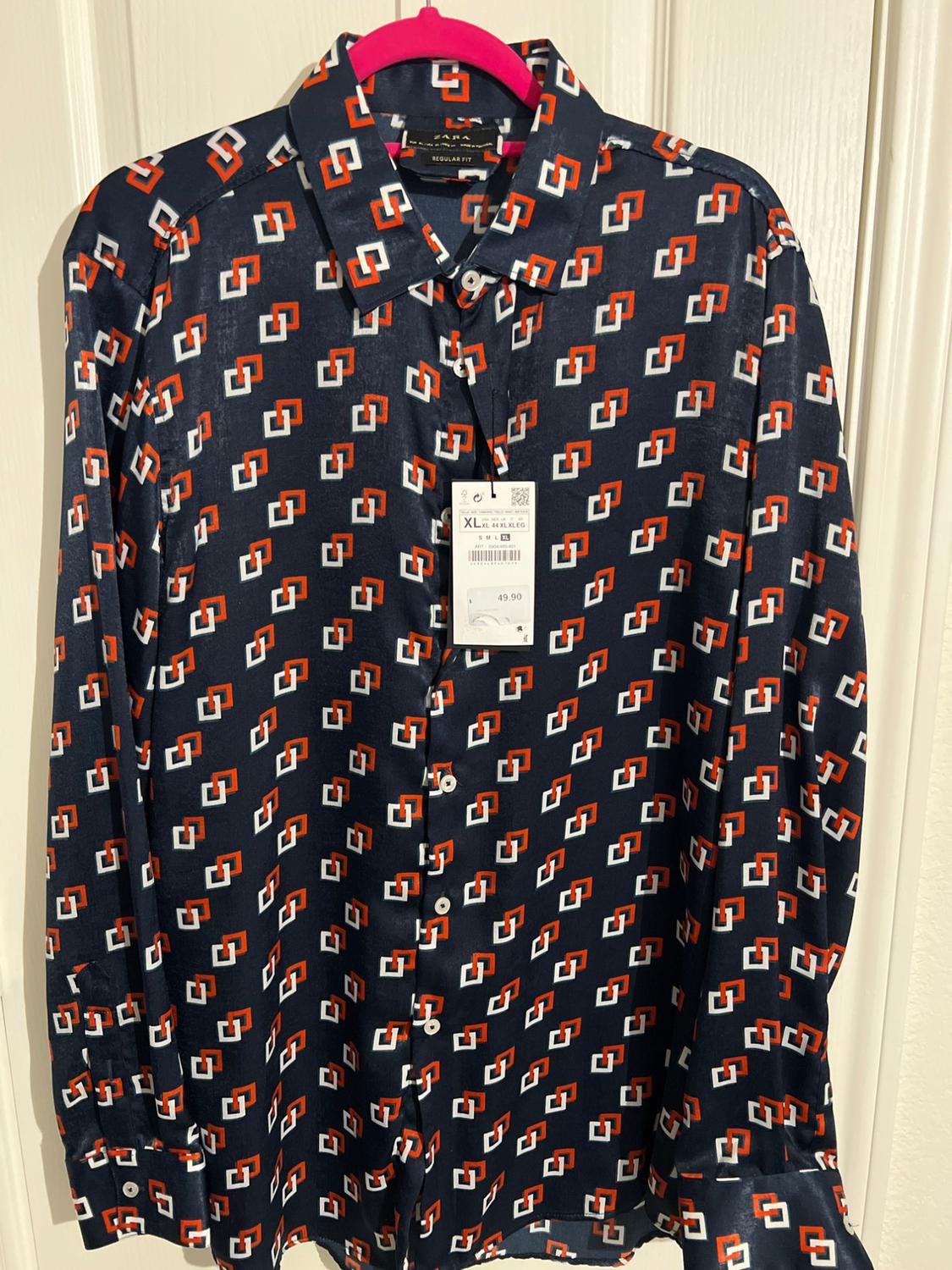 Long-sleeved shirt with geometric print in blue, red, and white, hanging on a pink hanger. Price tag visible.