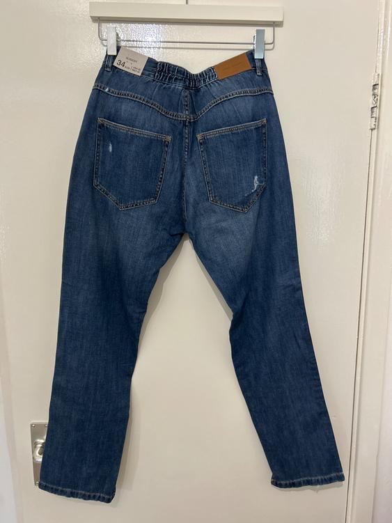 Dark blue jeans hanging on a white hanger, viewed from behind. Visible label: "Skinny 34". White door background.