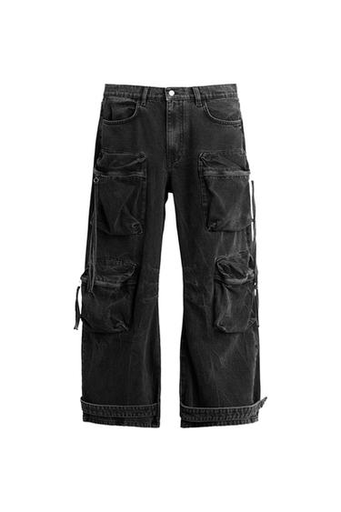 Black wide-leg cargo jeans with multiple pockets, dangling straps, and a hemmed cuff, on a white background.