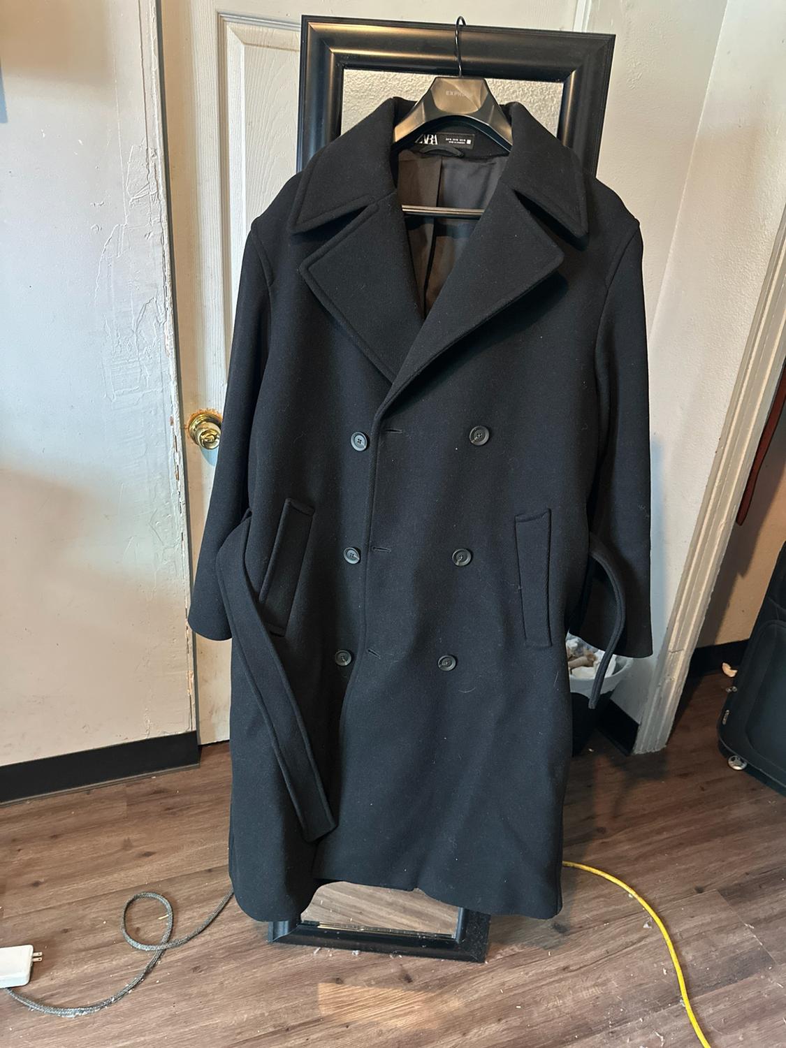 Black double-breasted overcoat with a belt and a zara label, hanging on a hanger above a mirror with a black frame in a room.