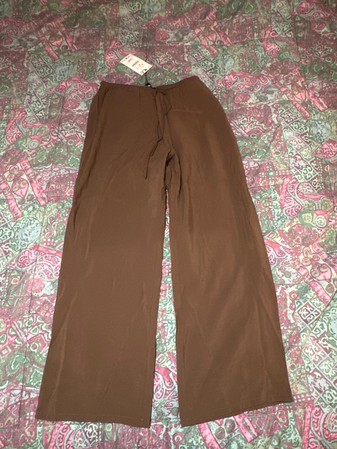 Brown wide-leg trousers with an elasticated drawstring waist, on a colourful patterned bedspread. Visible tag.