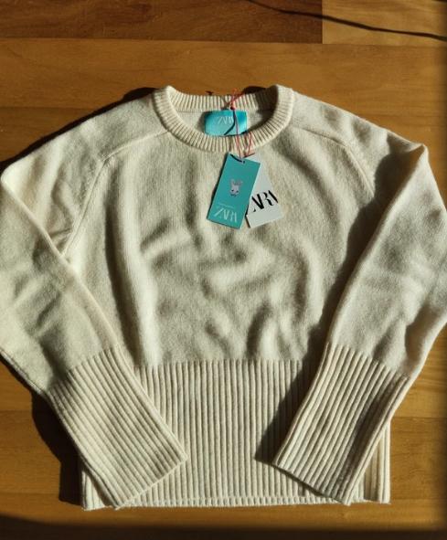White knitted jumper with hanging tags on a wooden surface. The tags show the text "ZVN".