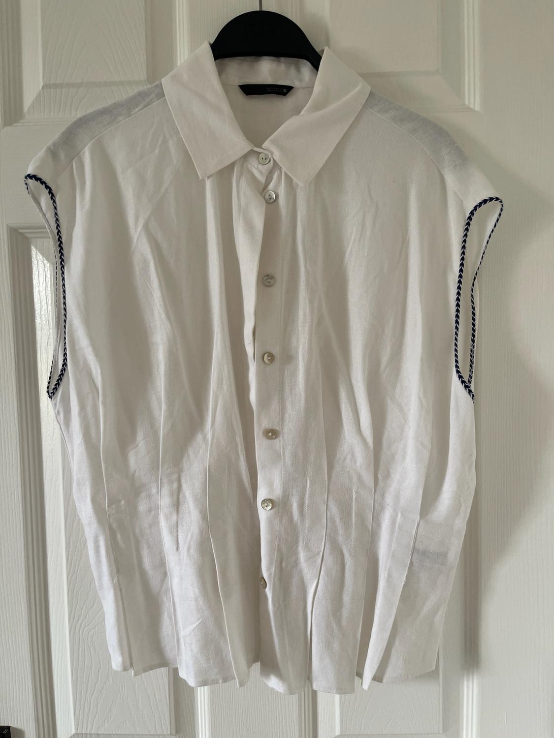 Sleeveless white shirt with a collar, front buttons, pleats and dark detailing at the armholes, hanging on a white door.