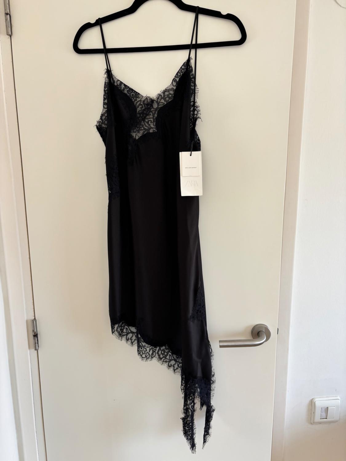 Black slip dress with lace and an asymmetrical hem, hanging on a hanger on a white door. Label with ZARA text.