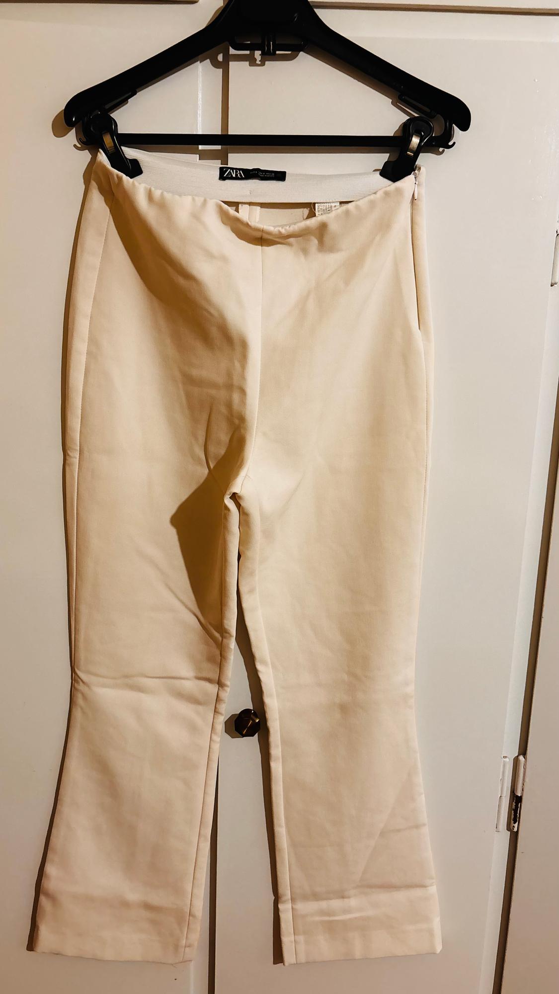 Beige trousers hanging on a black hanger over a white door. Visible tag with the brand "Zara".