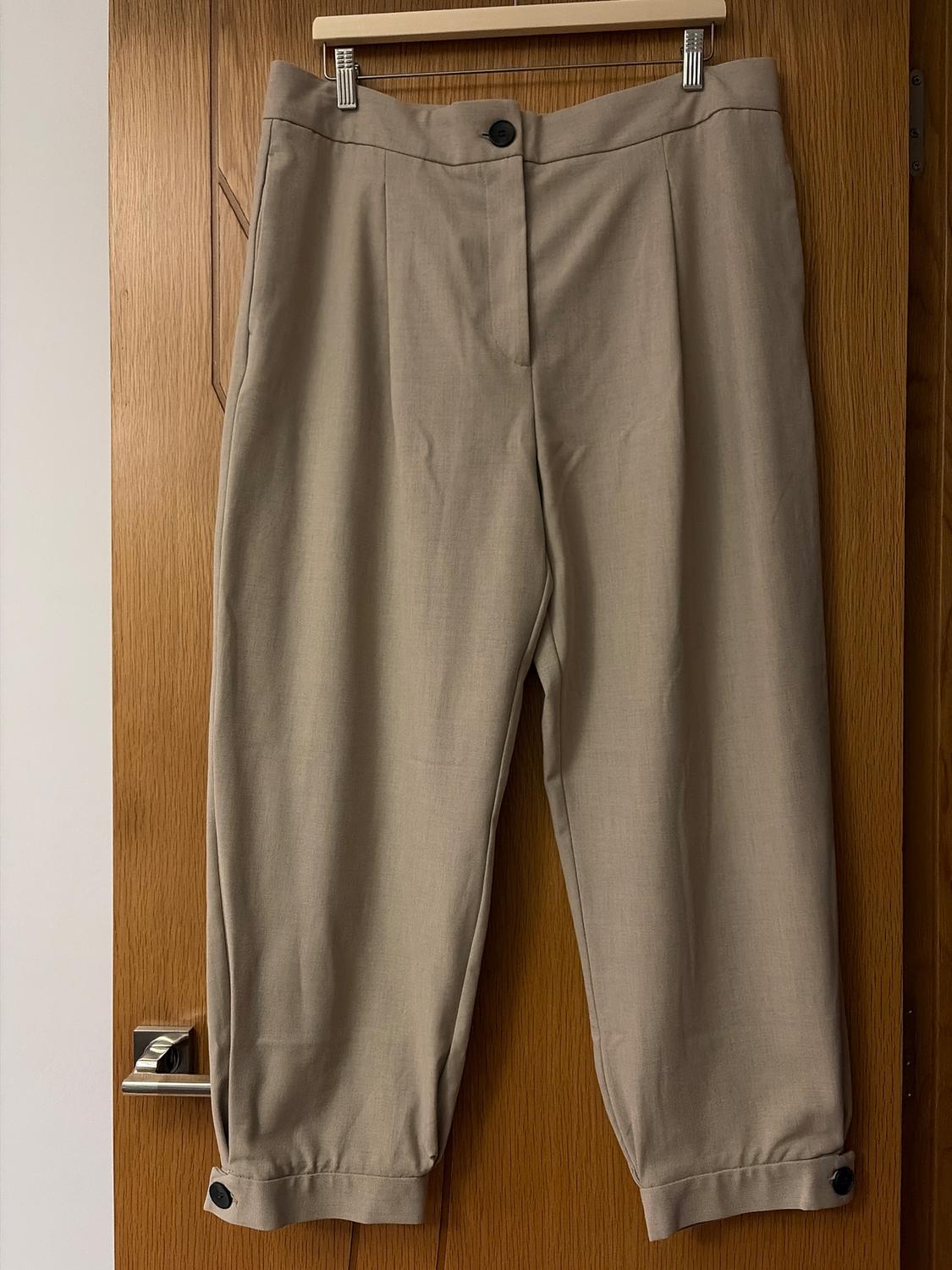 Beige trousers hanging on a wooden hanger over a wooden door. They have a black button at the waist and ankles.