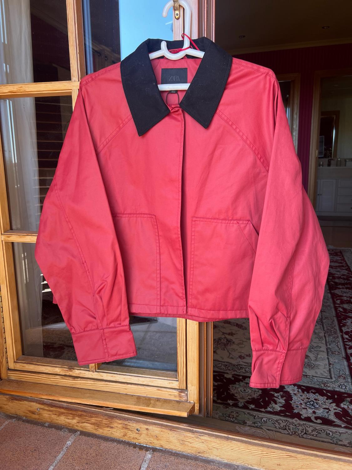 Red jacket with a black collar on a white hanger, hanging on a wooden door. ZARA label. Home setting with tiles and a rug.