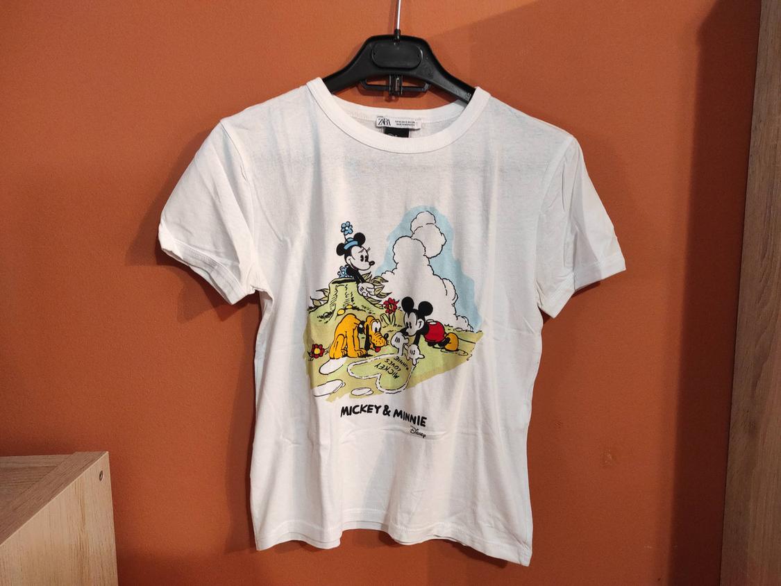 White T-shirt with Mickey and Minnie print in a landscape, hanging on a black hanger against an orange wall. Text: "MICKEY & MINNIE".