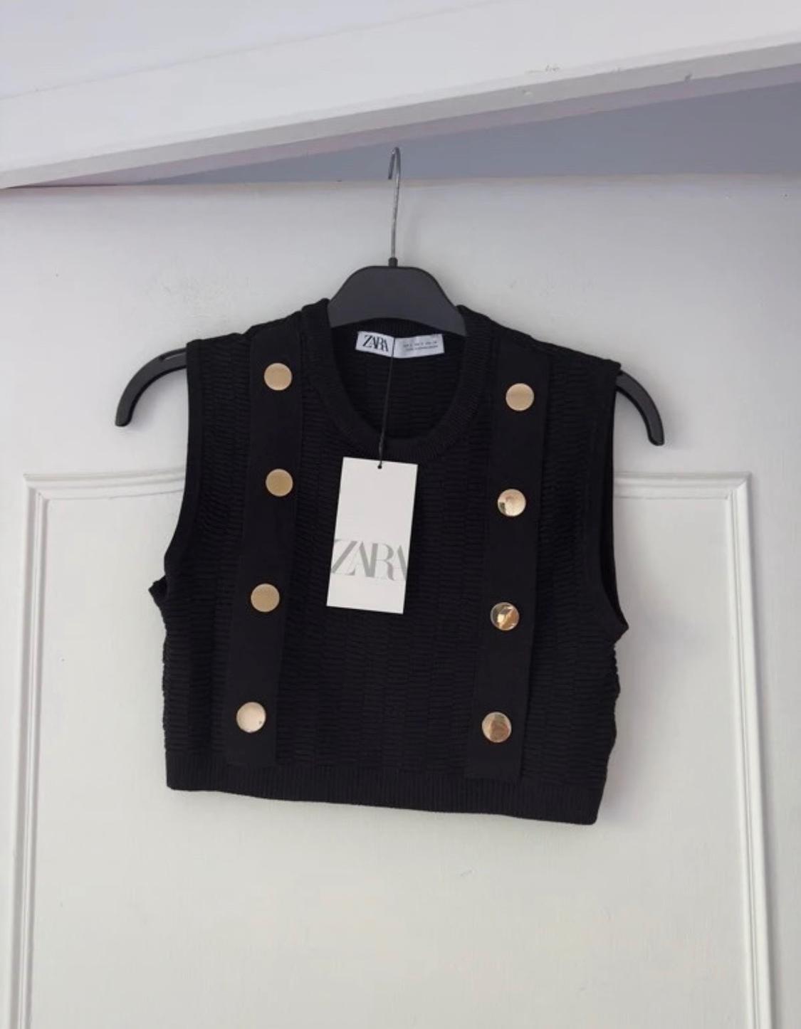 Black sleeveless top with gold buttons on the straps, hanging on a black hanger. Label with text "ZARA".