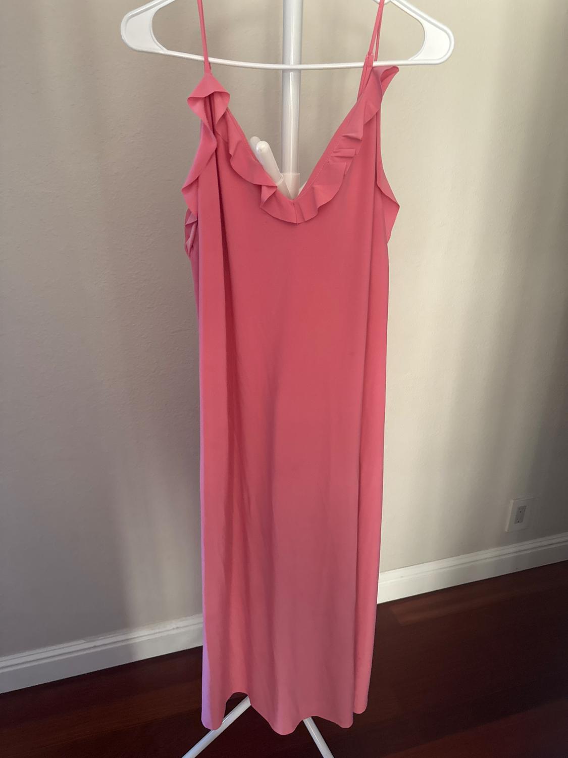 Long pink dress with ruffles on the neckline, hanging on a white hanger on a standing rack in front of a light wall.