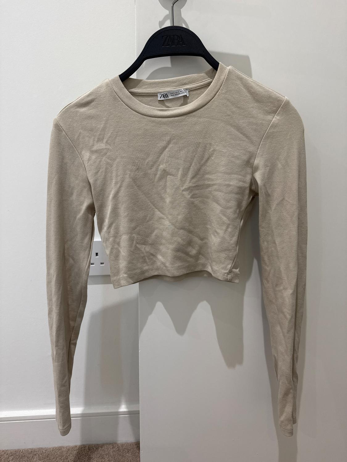 Long-sleeved, cropped beige t-shirt hanging on a black hanger with visible branding, on a white background.