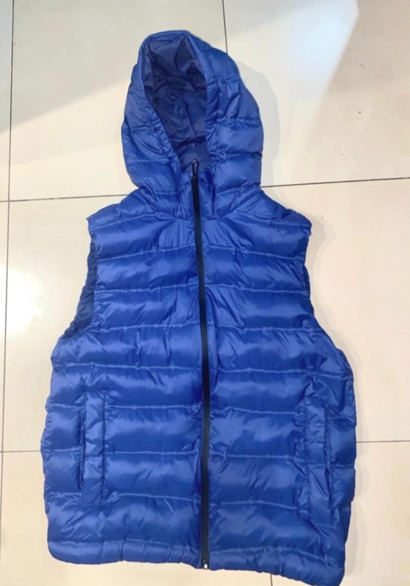 Blue padded gilet with hood and front zip, placed on a light tiled floor.