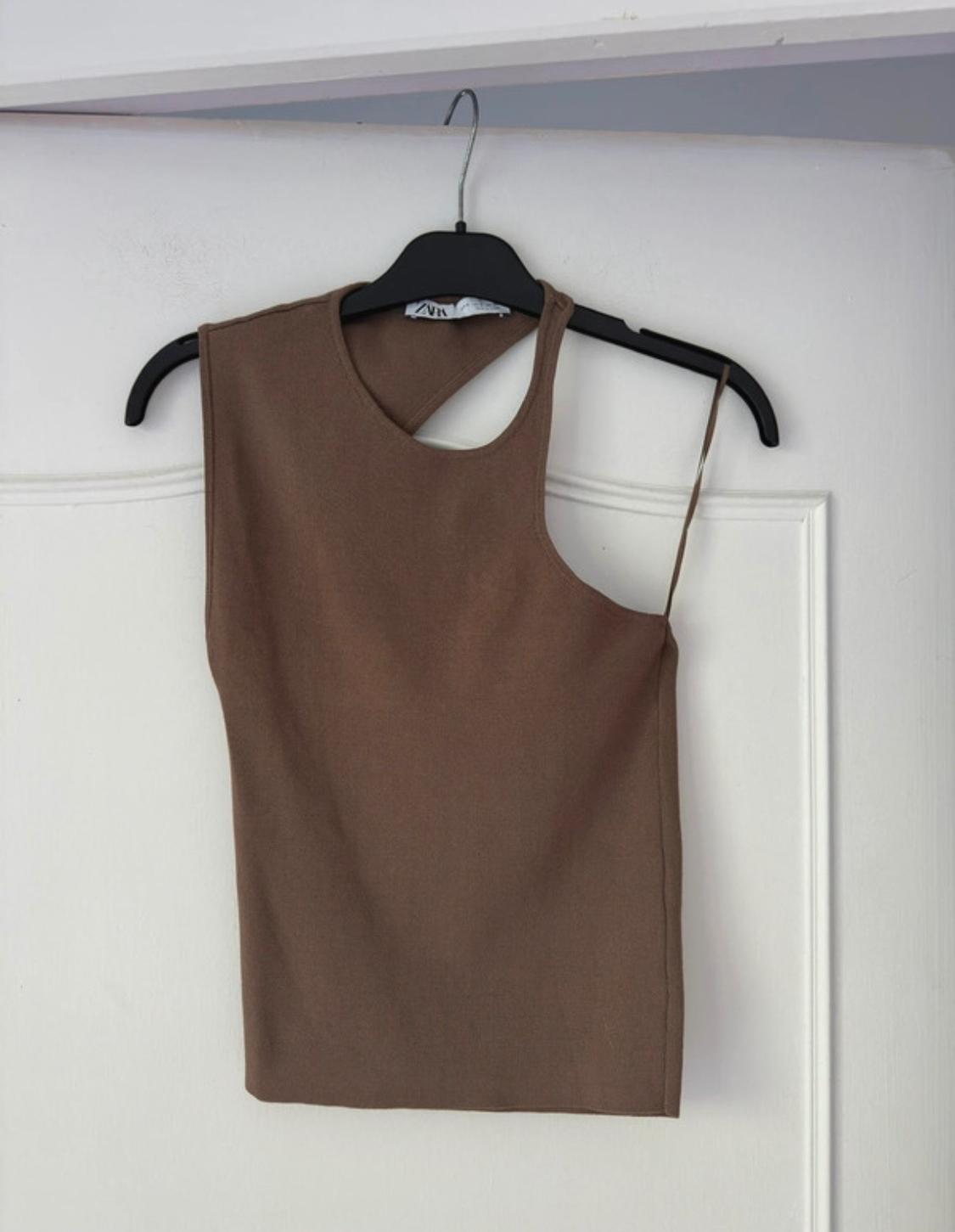 Brown sleeveless top with an asymmetrical design, hanging on a black hanger over a white door.