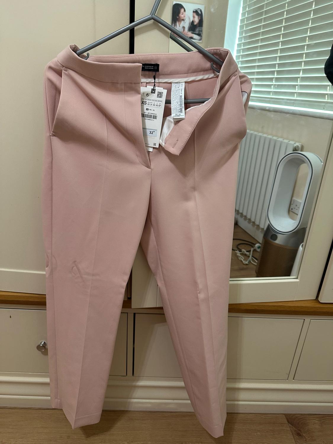 Light pink trousers hanging on a hanger in front of a mirror, with visible tags. Room background with blinds and radiator.