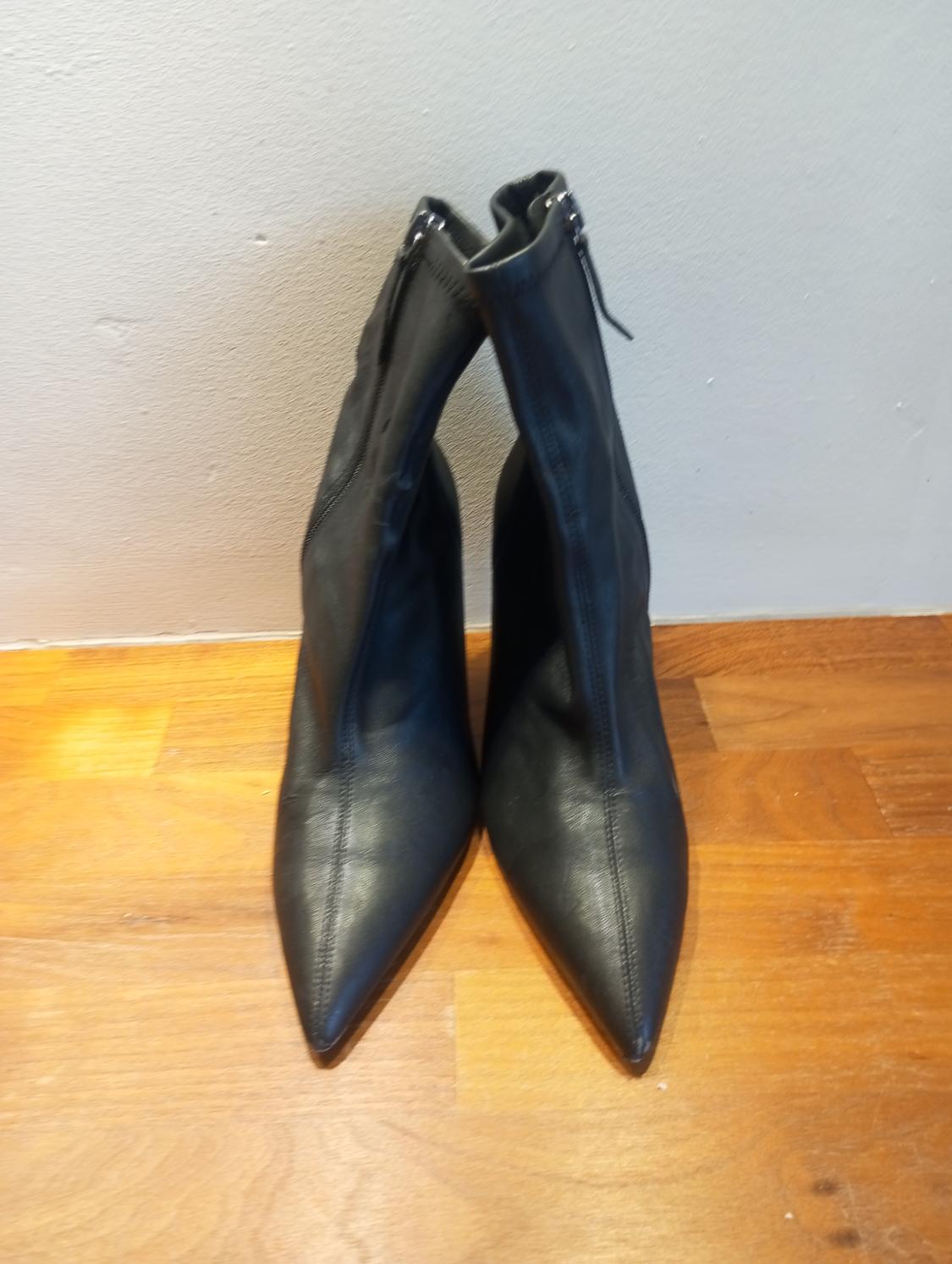 Black pointed-toe boots with a zip, placed on a light wooden floor, white wall background.