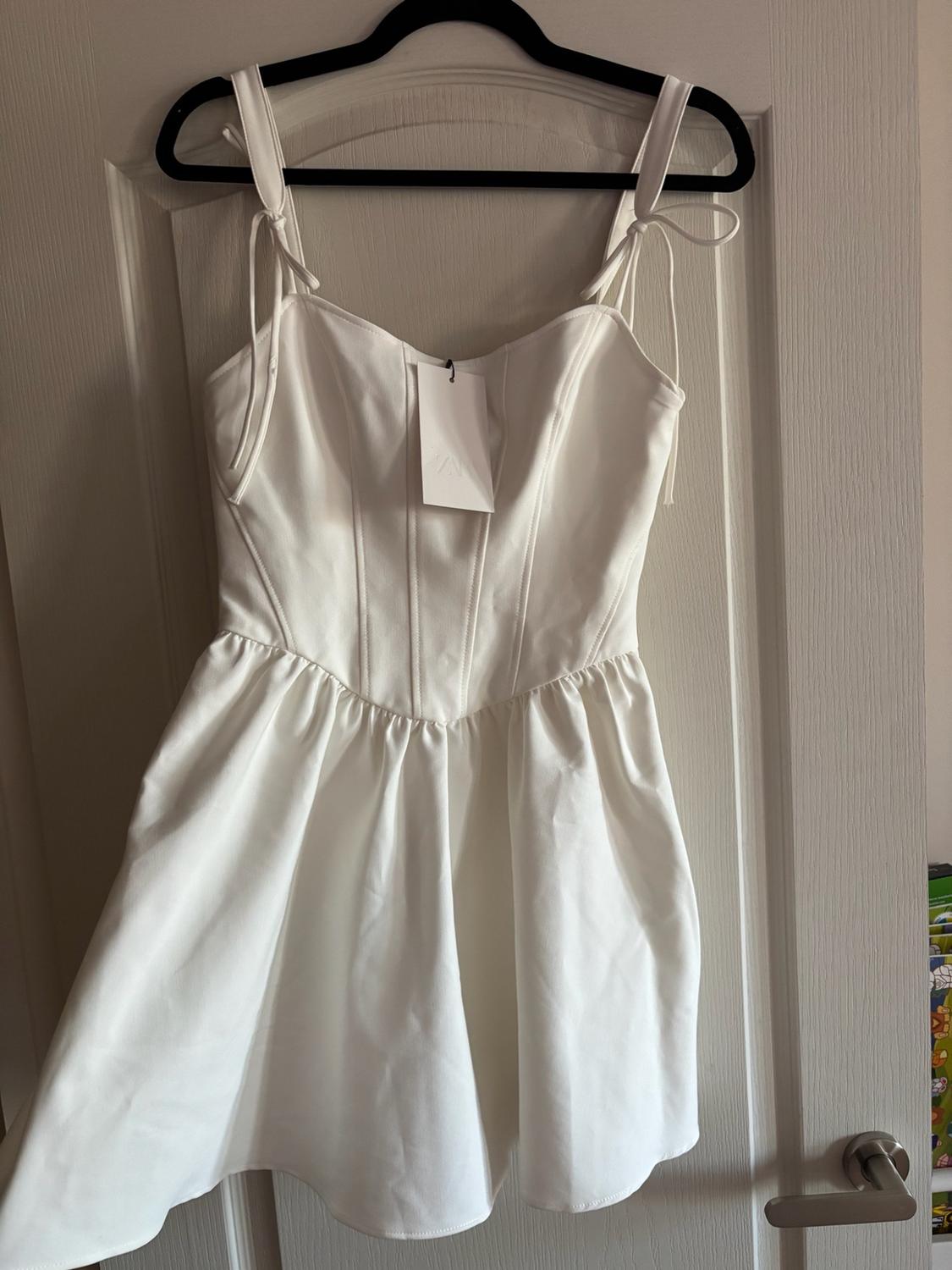 White short dress with tags, ribbon straps, corset-style bodice and flared skirt, hanging on a black hanger over a door.