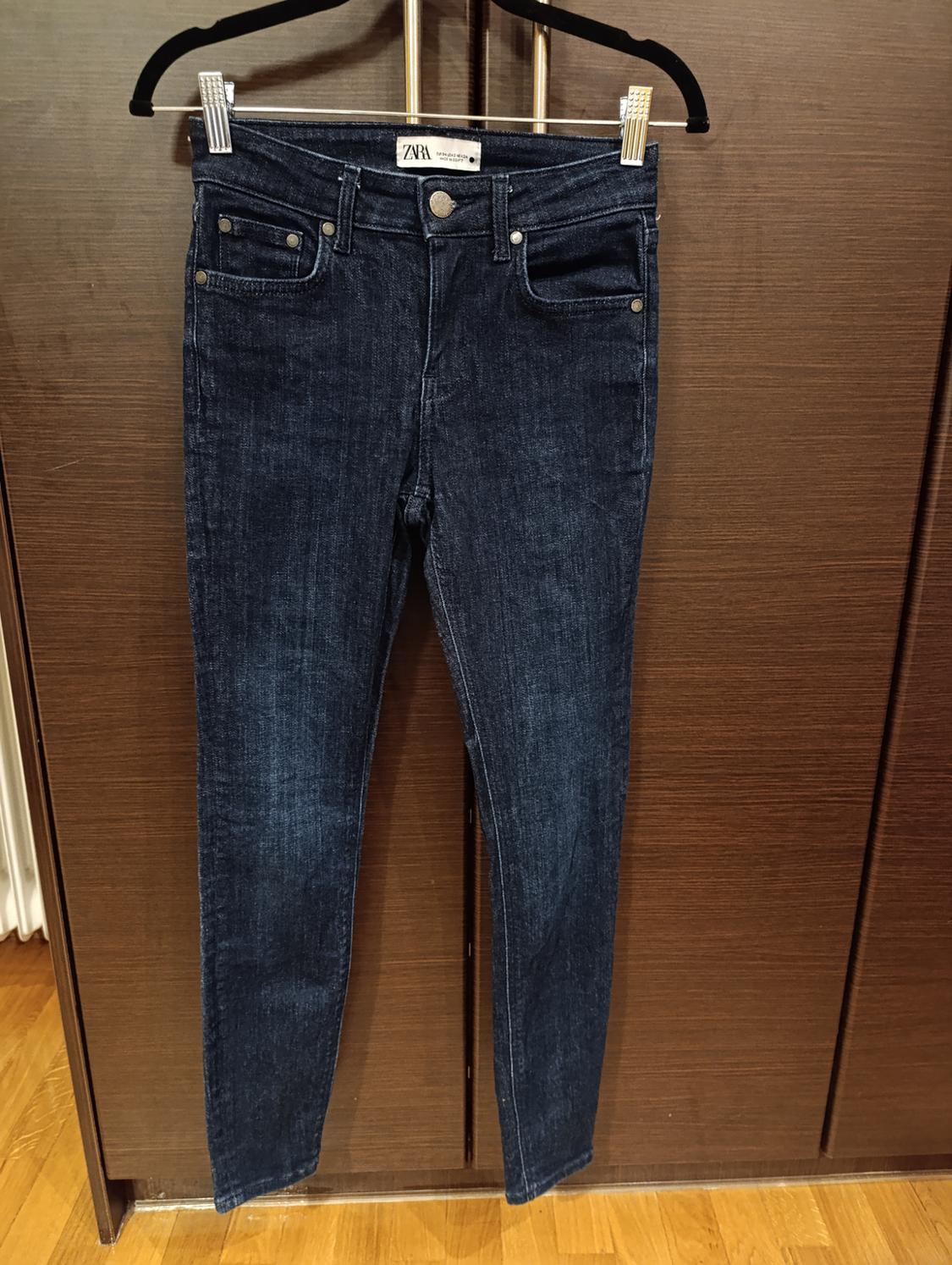 Dark blue jeans hanging on a black hanger in front of a wooden background. Visible label with the text "ZARA".