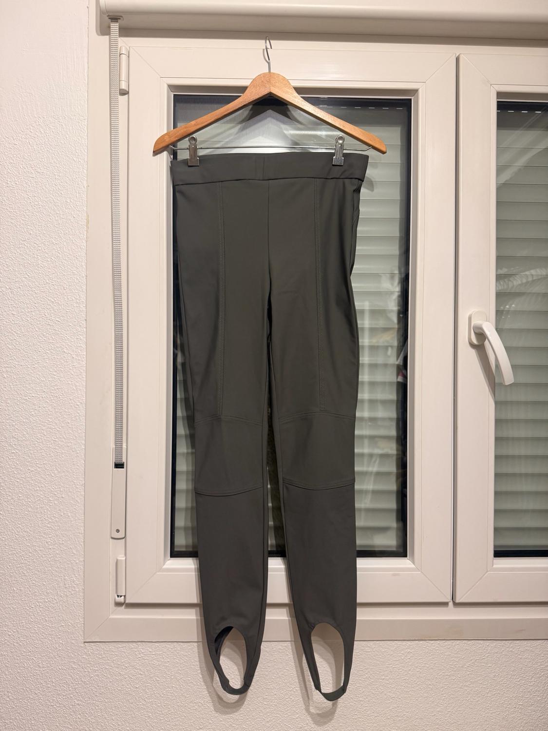 Dark green stirrup trousers with seams, hanging on a wooden hanger in front of a white window.