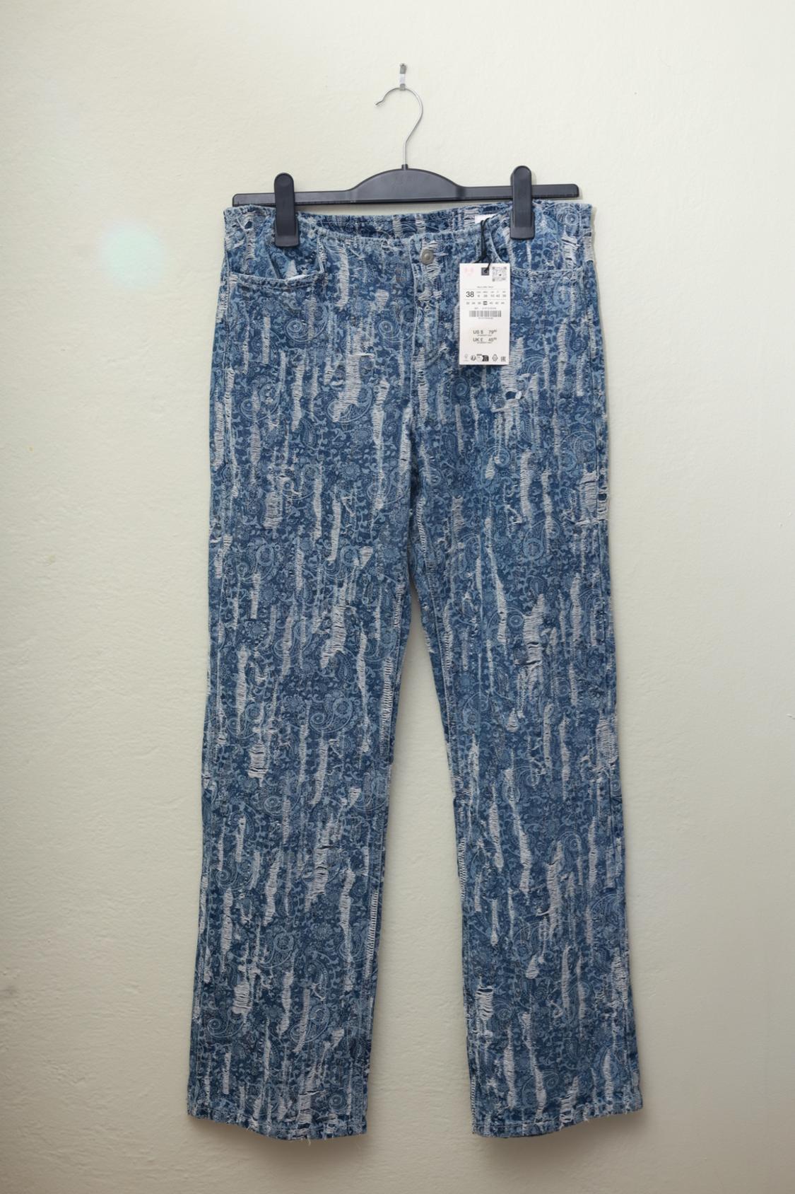 Blue jeans with a print and vertical distressed details, hanging from a black hanger against a light wall.