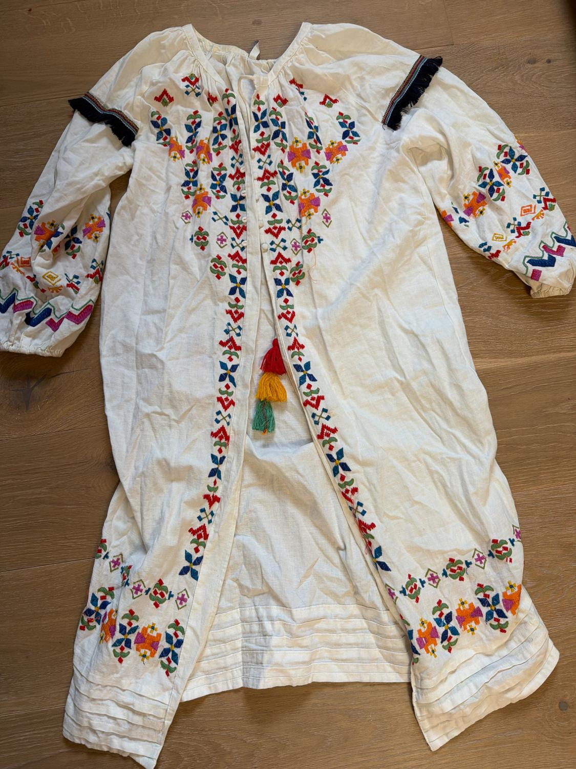 White dress with colourful geometric and floral embroidery, with tassels on the front, on a wooden floor.