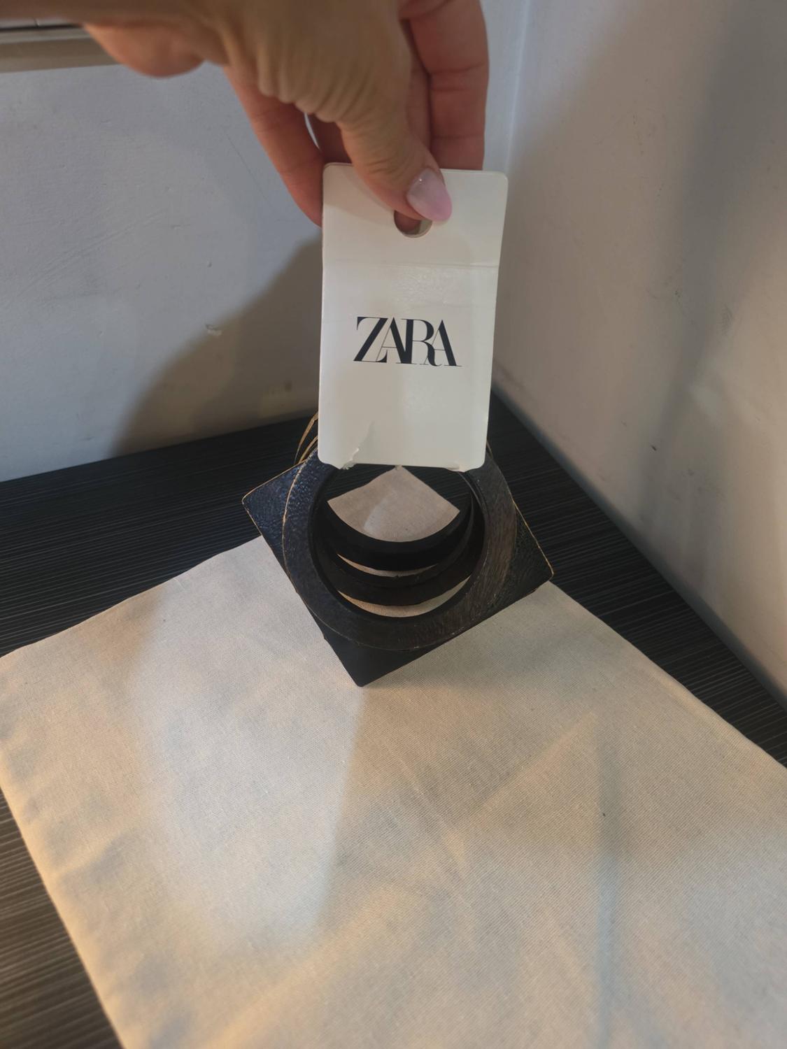 Hand holding a tag with the text ZARA over dark square bracelets resting on a white fabric.