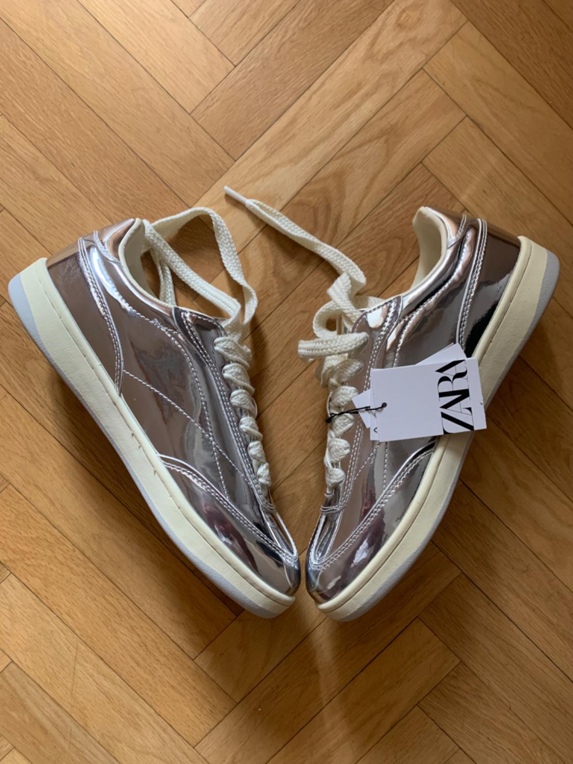 Metallic silver trainers with white laces on a wooden floor. Label with the text ZARA.
