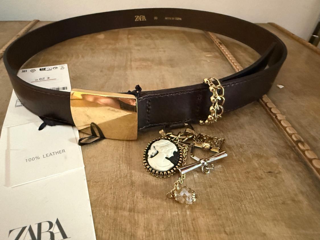 Brown belt with gold buckle and decorative charms on a wooden table. Visible text: ZARA, 100% LEATHER.