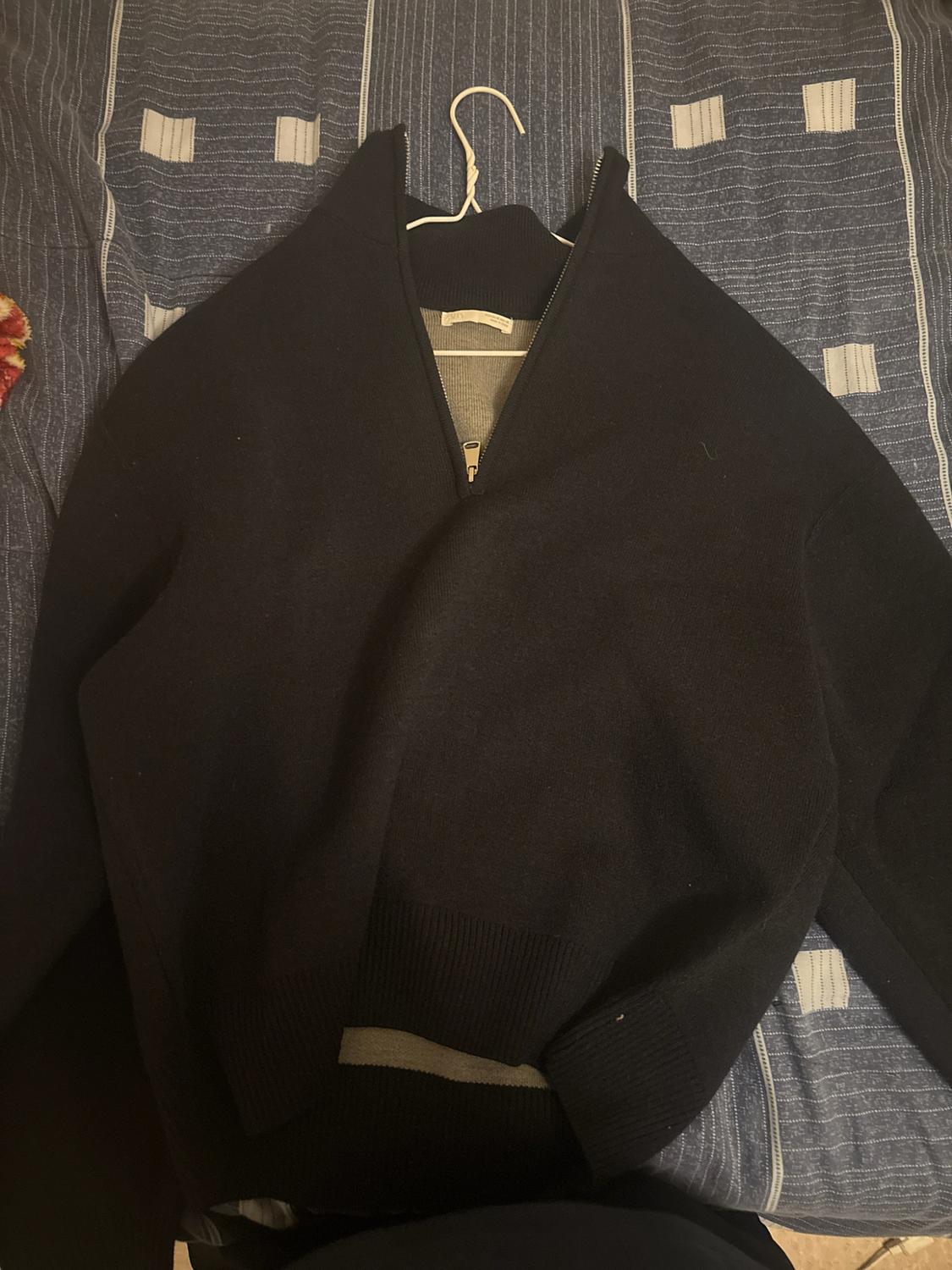 Dark blue knit zip-up with a front zipper, a high neck, and ribbed cuffs, hanging on a hanger over a blue striped quilt.