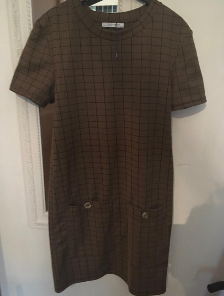 Brown checked dress with short sleeves and two front pockets with buttons, hanging on a hanger against a light background.