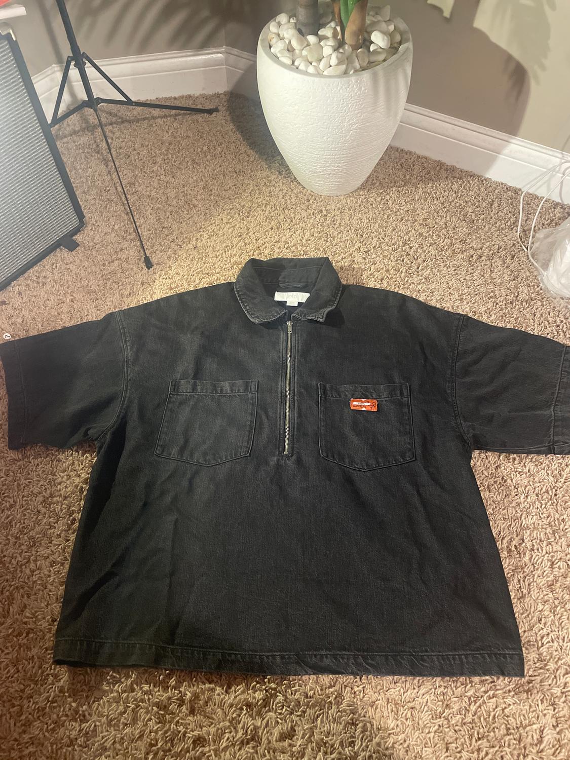 Black short-sleeved shirt with zip and two front pockets, on a beige rug. Potted plant in a white pot in the background.